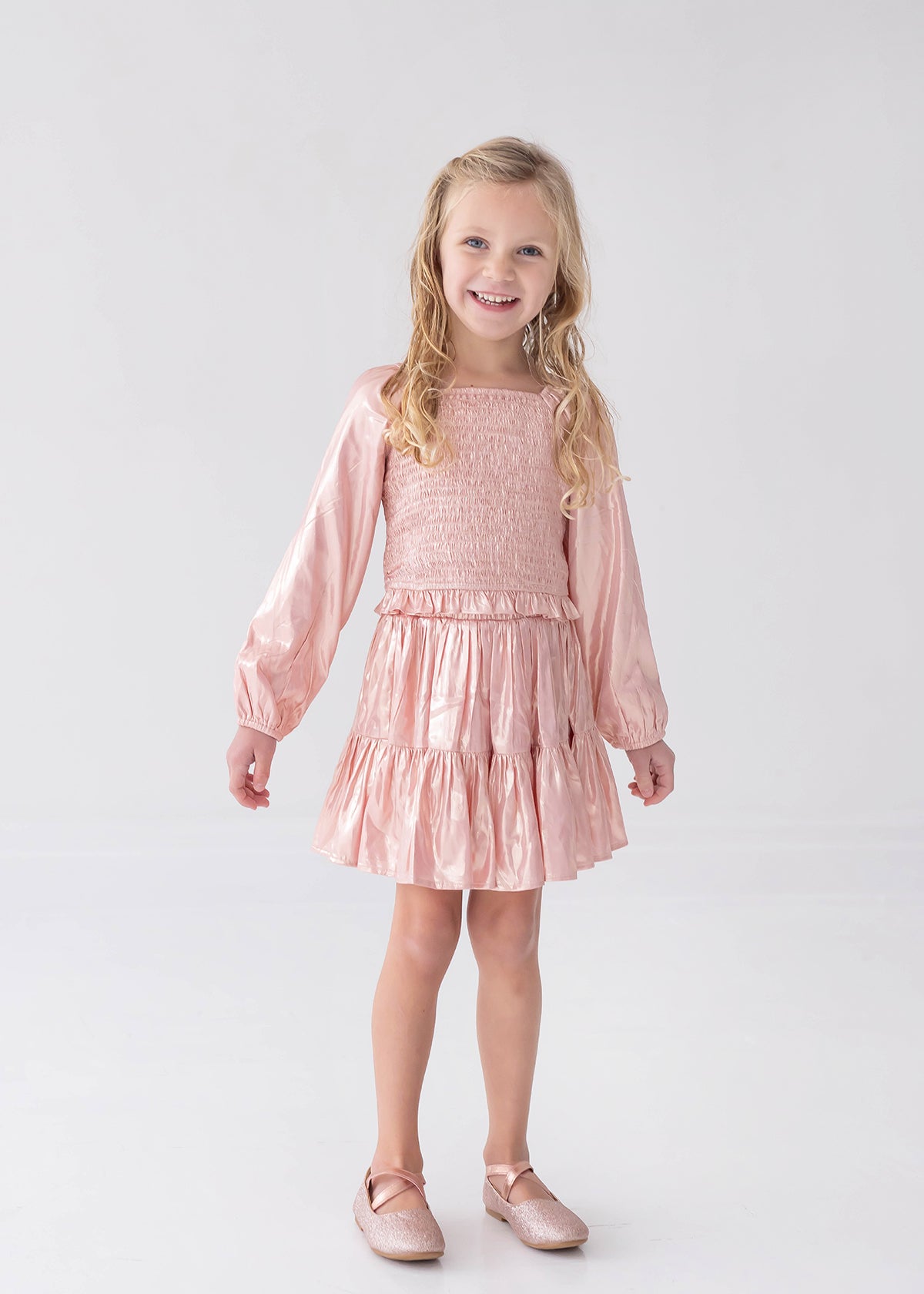 A young girl with long blonde hair wears the Rosy Glow Long Sleeve Smocked Metallic Tiered Skirt Set by Isobella and Chloe, featuring a shiny light pink crop top and matching skirt, paired with coordinating pink shoes, standing and smiling against a plain white background—an adorable special occasion outfit.