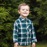 A young boy with short brown hair smiles while standing outdoors, wearing the Beckett and Bear Christopher Long Sleeve Plaid Button Down Chest Pocket Shirt and tan pants, surrounded by lush green foliage.