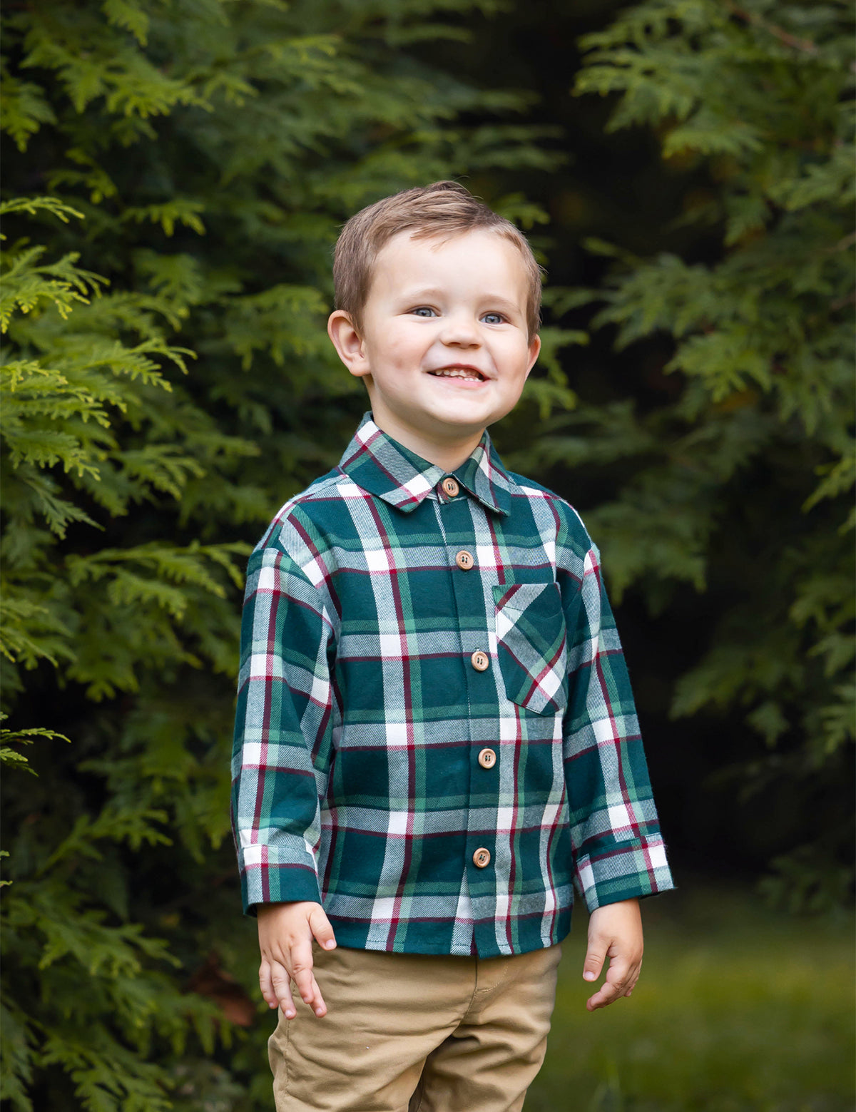 A young boy with short brown hair smiles while standing outdoors, wearing the Beckett and Bear Christopher Long Sleeve Plaid Button Down Chest Pocket Shirt and tan pants, surrounded by lush green foliage.