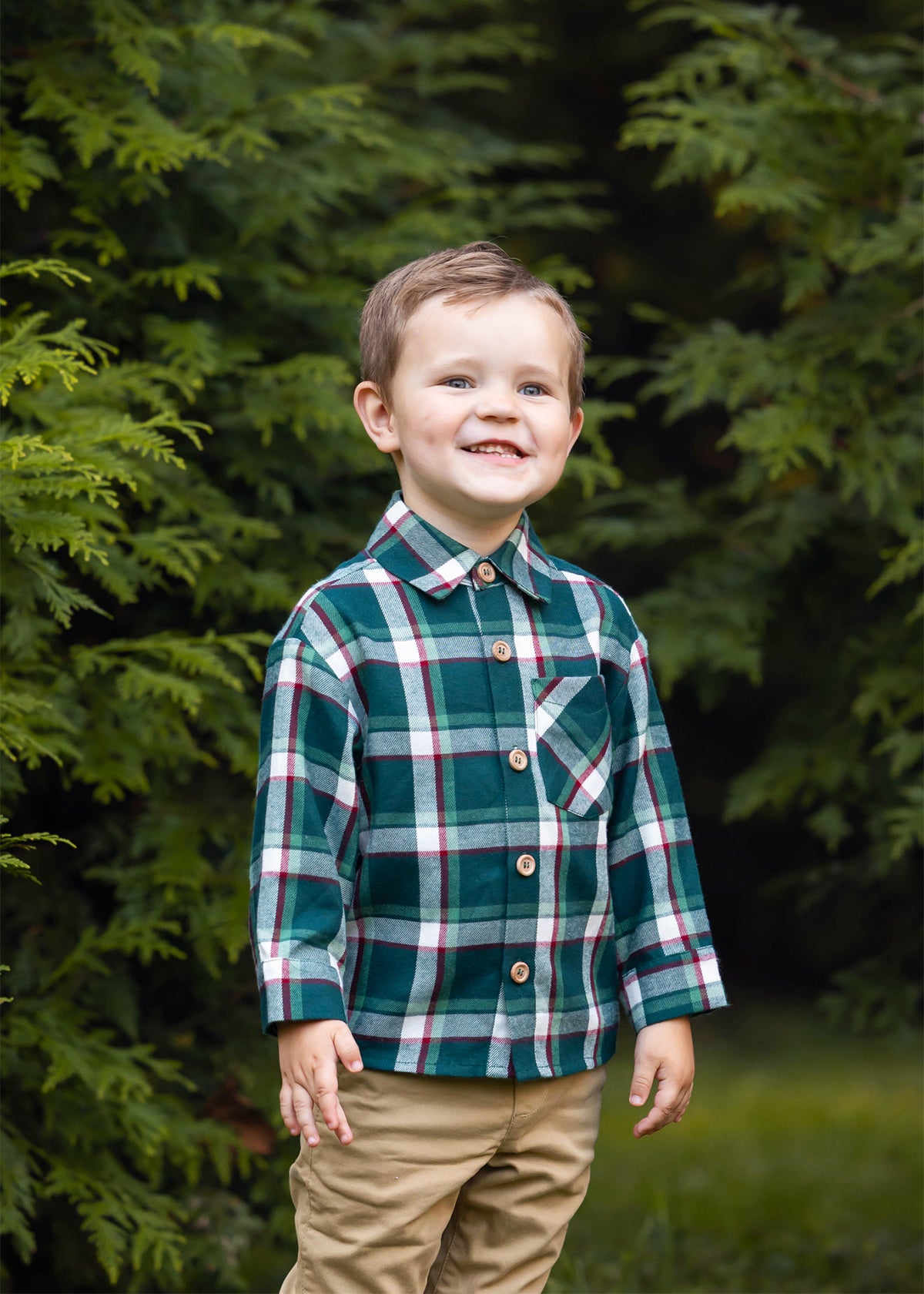 A young boy with short brown hair smiles while standing outdoors, wearing the Beckett and Bear Christopher Long Sleeve Plaid Button Down Chest Pocket Shirt and tan pants, surrounded by lush green foliage.