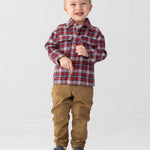 A smiling young boy with short hair wears the Beckett and Bear Everett Long Sleeve Plaid Flannel Button Front Chest Pocket Shirt in red, paired with tan pants and gray sneakers, standing on a wooden box against a plain light background.
