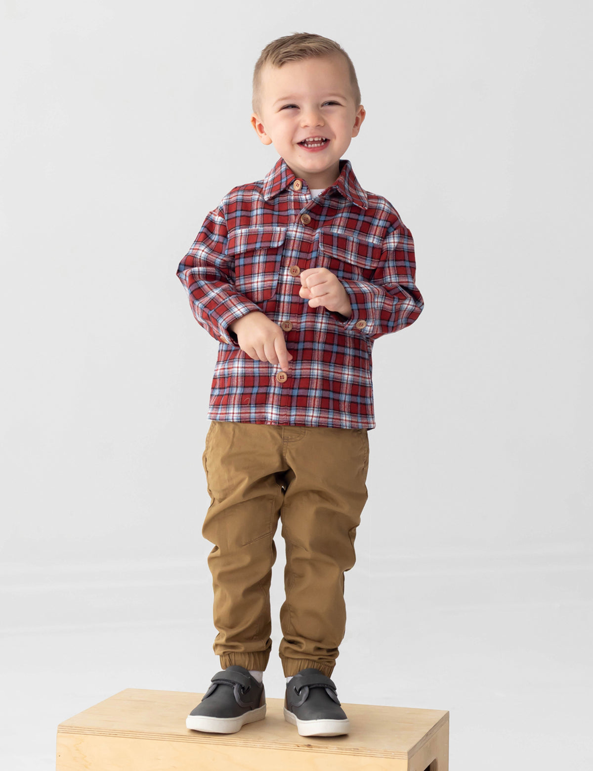 A smiling young boy with short hair wears the Beckett and Bear Everett Long Sleeve Plaid Flannel Button Front Chest Pocket Shirt in red, paired with tan pants and gray sneakers, standing on a wooden box against a plain light background.