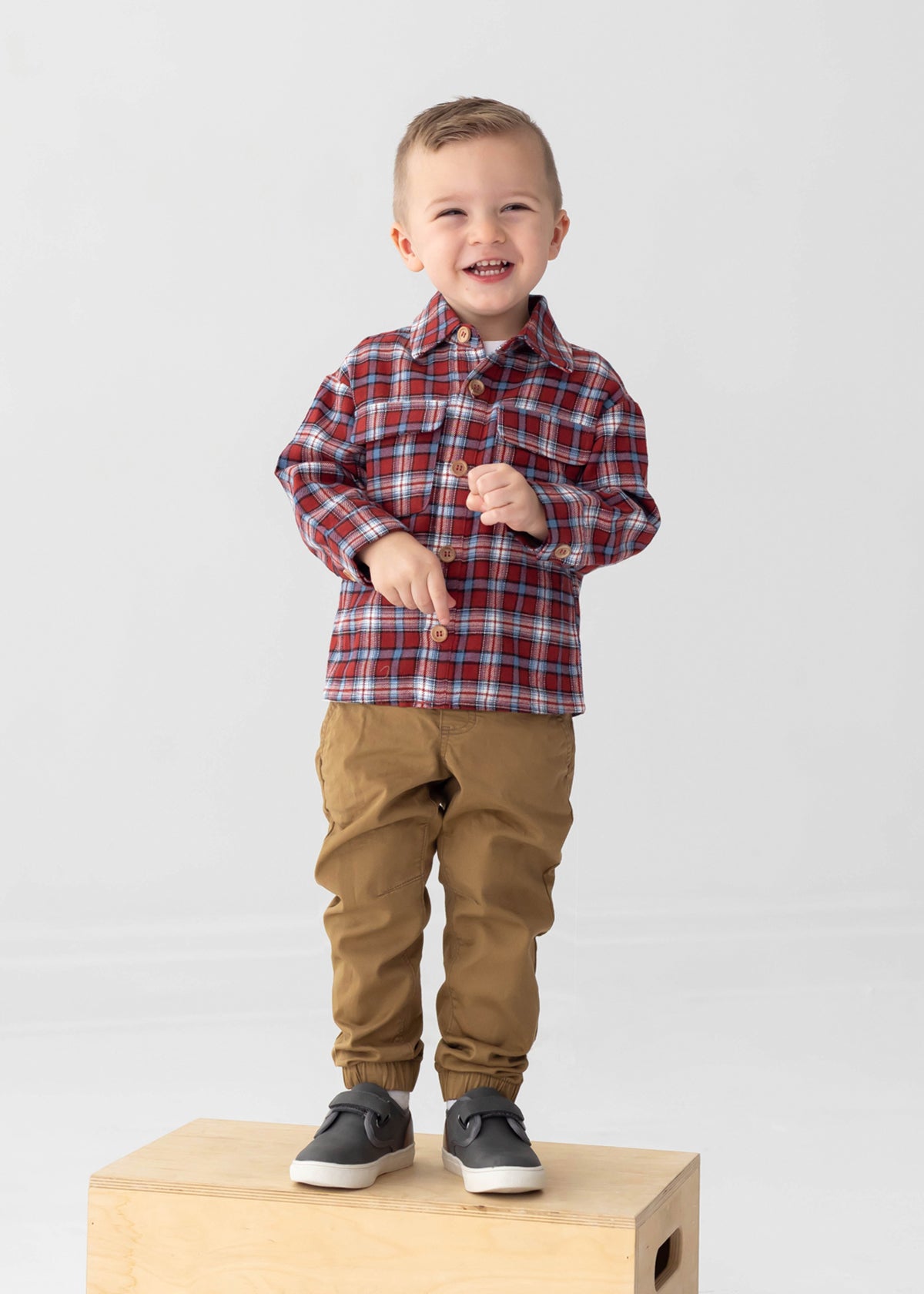 A smiling young boy with short hair wears the Beckett and Bear Everett Long Sleeve Plaid Flannel Button Front Chest Pocket Shirt in red, paired with tan pants and gray sneakers, standing on a wooden box against a plain light background.