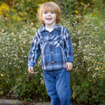 A young child with light brown hair wears the Beckett and Bear Hunter Long Sleeve Plaid Flannel Chest Pocket Shirt, paired with jeans and brown shoes, smiling while standing in front of green foliage and small white flowers on a sunny day.
