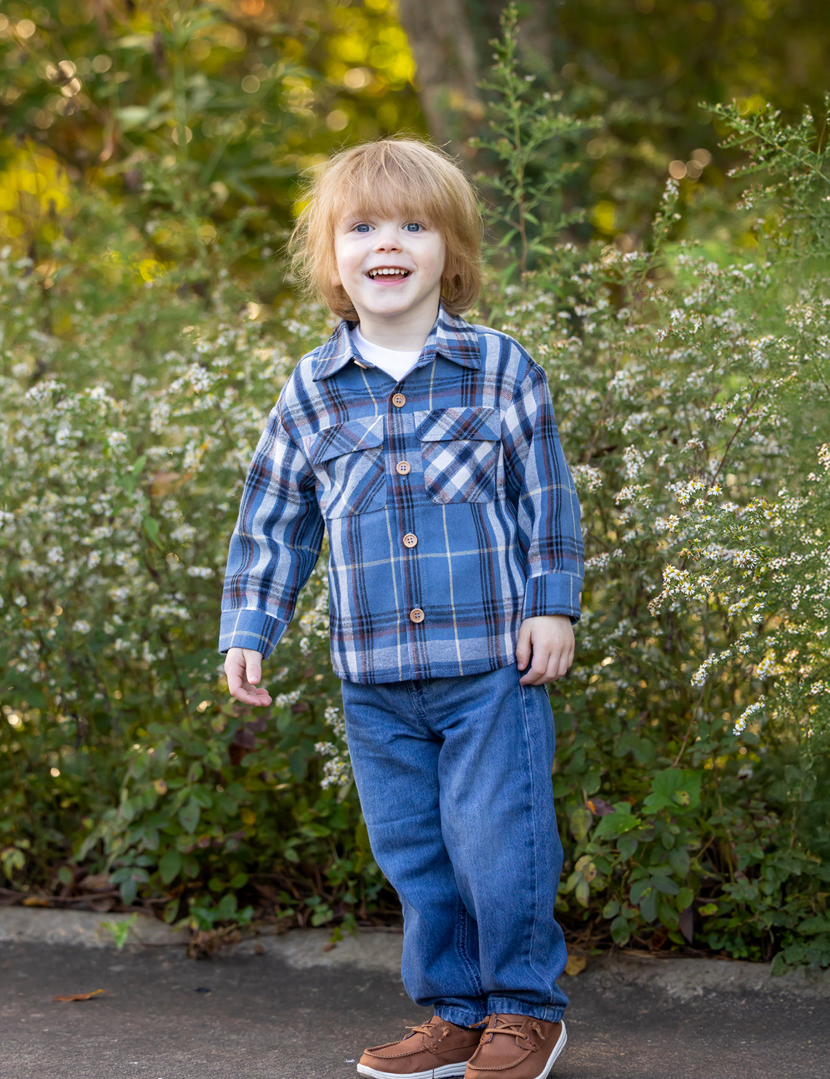 A young child with light brown hair wears the Beckett and Bear Hunter Long Sleeve Plaid Flannel Chest Pocket Shirt, paired with jeans and brown shoes, smiling while standing in front of green foliage and small white flowers on a sunny day.
