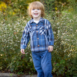 A young child with light brown hair wears the Beckett and Bear Hunter Long Sleeve Plaid Flannel Chest Pocket Shirt, paired with jeans and brown shoes, smiling while standing in front of green foliage and small white flowers on a sunny day.