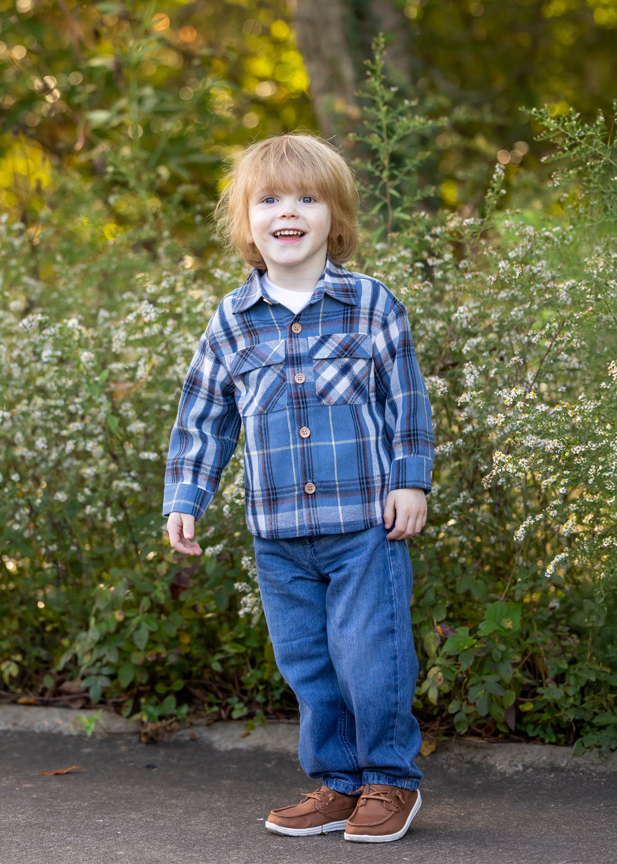 A young child with light brown hair wears the Beckett and Bear Hunter Long Sleeve Plaid Flannel Chest Pocket Shirt, paired with jeans and brown shoes, smiling while standing in front of green foliage and small white flowers on a sunny day.