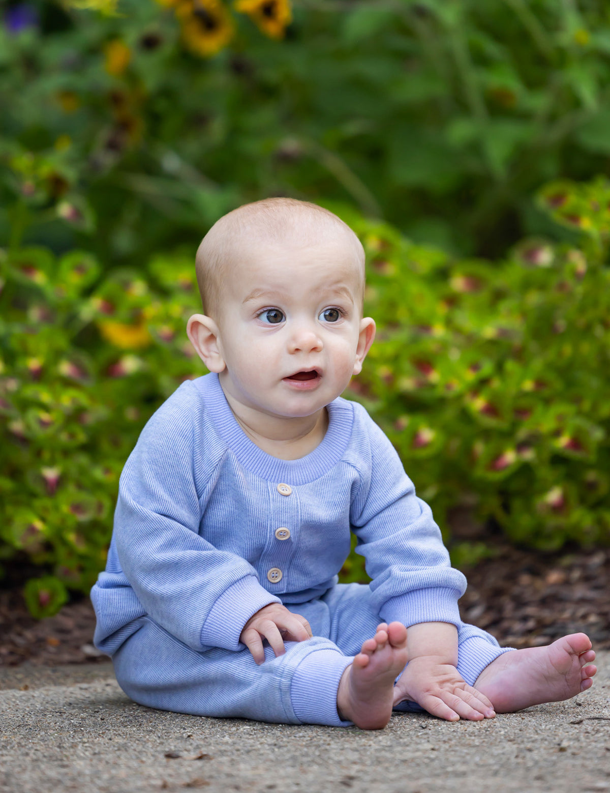 A baby wearing the Beckett and Bear Daniel Long Sleeve Solid Ribbed Button Placket Straight Leg Romper in heathered blue sits on a path surrounded by green plants and yellow flowers, gazing upward with a curious expression.