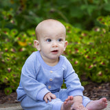 A baby wearing the Beckett and Bear Daniel Long Sleeve Solid Ribbed Button Placket Straight Leg Romper in heathered blue sits on a path surrounded by green plants and yellow flowers, gazing upward with a curious expression.