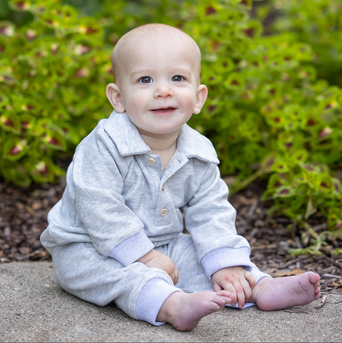 A baby in the Beckett and Bear Jacob Long Sleeve Solid Collared Ribbed Trim Romper, crafted from a soft cotton-blend knit in heathered gray, sits barefoot on a sidewalk and smiles at the camera with green plants in the background. Snap closures offer added convenience to this charming outfit.
