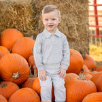 A young child wearing the Beckett and Bear Jacob Long Sleeve Solid Collared Sweatshirt and Jogger Set in soft brushed knit heather gray stands among orange pumpkins, smiling at the camera. The matching elastic waist joggers and collared sweatshirt provide both comfort and style. Hay bales and more pumpkins in the background evoke a cozy pumpkin patch scene.