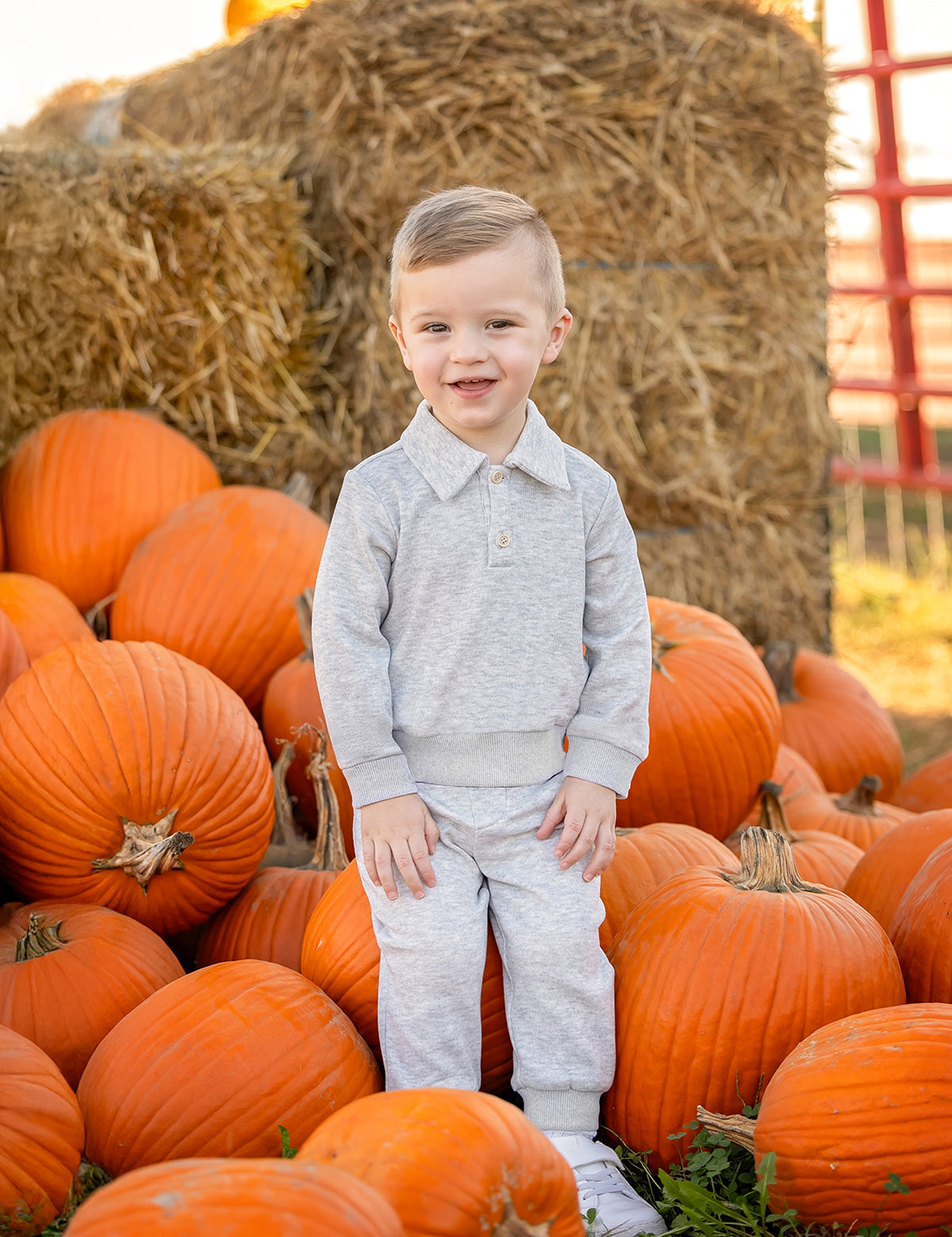 A young child wearing the Beckett and Bear Jacob Long Sleeve Solid Collared Sweatshirt and Jogger Set in soft brushed knit heather gray stands among orange pumpkins, smiling at the camera. The matching elastic waist joggers and collared sweatshirt provide both comfort and style. Hay bales and more pumpkins in the background evoke a cozy pumpkin patch scene.