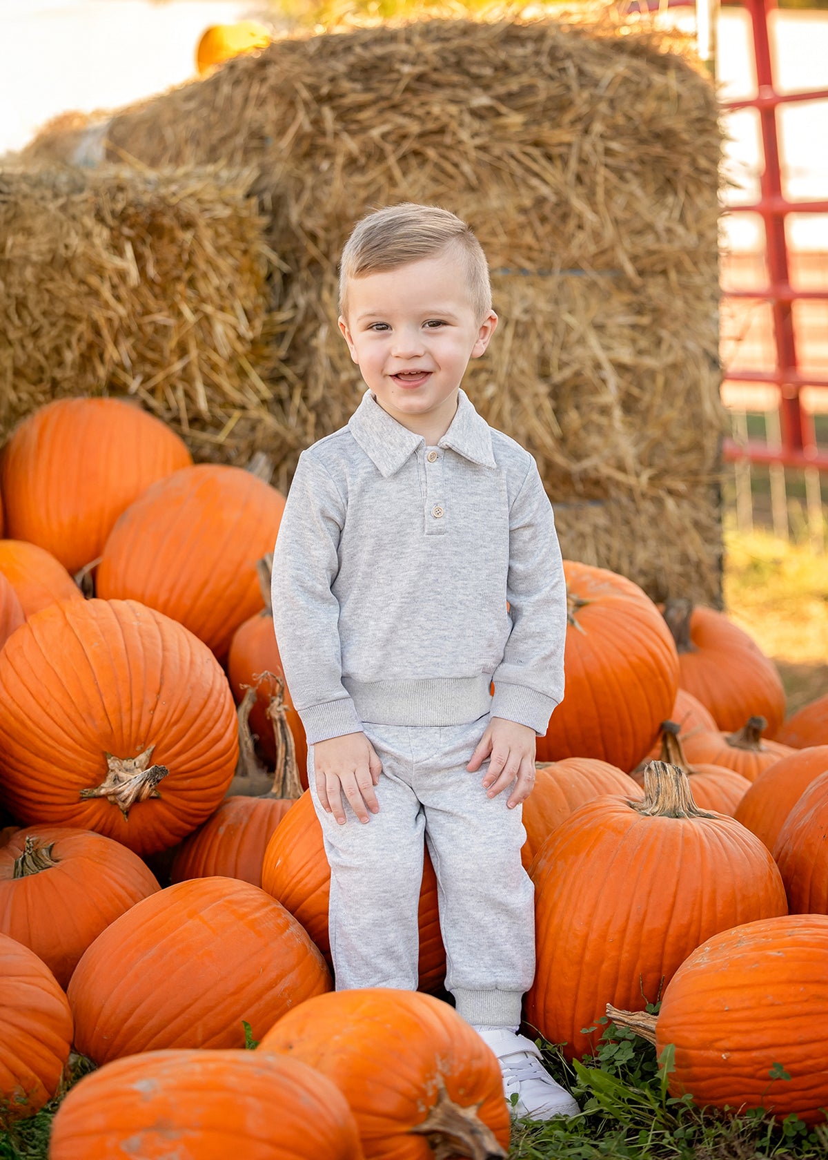 A young child wearing the Beckett and Bear Jacob Long Sleeve Solid Collared Sweatshirt and Jogger Set in soft brushed knit heather gray stands among orange pumpkins, smiling at the camera. The matching elastic waist joggers and collared sweatshirt provide both comfort and style. Hay bales and more pumpkins in the background evoke a cozy pumpkin patch scene.
