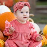 Dressed in the Mabel and Honey Poppy Rose Long Sleeve Solid Ribbed Bow Tie Bubble Romper with a matching headband, a baby sits among orange and white pumpkins outdoors, gazing slightly upward with a curious expression.