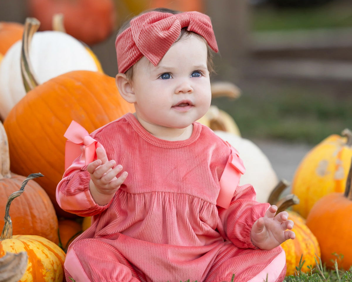 Dressed in the Mabel and Honey Poppy Rose Long Sleeve Solid Ribbed Bow Tie Bubble Romper with a matching headband, a baby sits among orange and white pumpkins outdoors, gazing slightly upward with a curious expression.