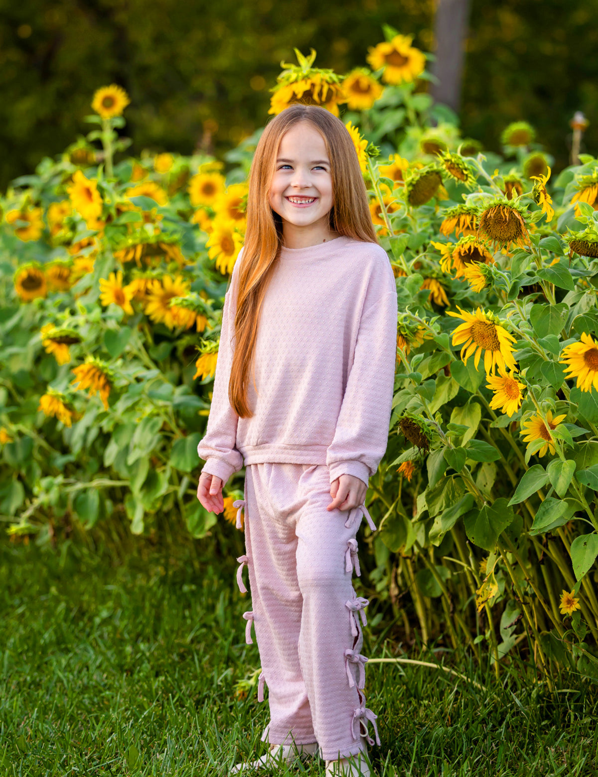 A young girl with long red hair smiles while standing in front of a field of blooming sunflowers. She wears the Mabel and Honey Emma Long Sleeve Textured Bow Tie Trim Top and Pant Set, featuring relaxed wide leg pants, and stands on green grass.