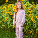 A young girl with long red hair smiles while standing in front of a field of blooming sunflowers. She wears the Mabel and Honey Emma Long Sleeve Textured Bow Tie Trim Top and Pant Set, featuring relaxed wide leg pants, and stands on green grass.