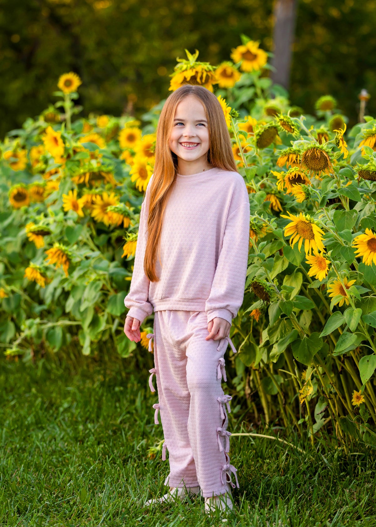 A young girl with long red hair smiles while standing in front of a field of blooming sunflowers. She wears the Mabel and Honey Emma Long Sleeve Textured Bow Tie Trim Top and Pant Set, featuring relaxed wide leg pants, and stands on green grass.