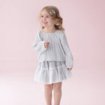 A young girl with blonde hair is wearing the Isobella and Chloe Shimmer Shine Long Sleeve Metallic Pleated Top and Tiered Skirt Set, styled with gray shoes, as she stands smiling against a pale pink background—an adorable choice for a girls party outfit.