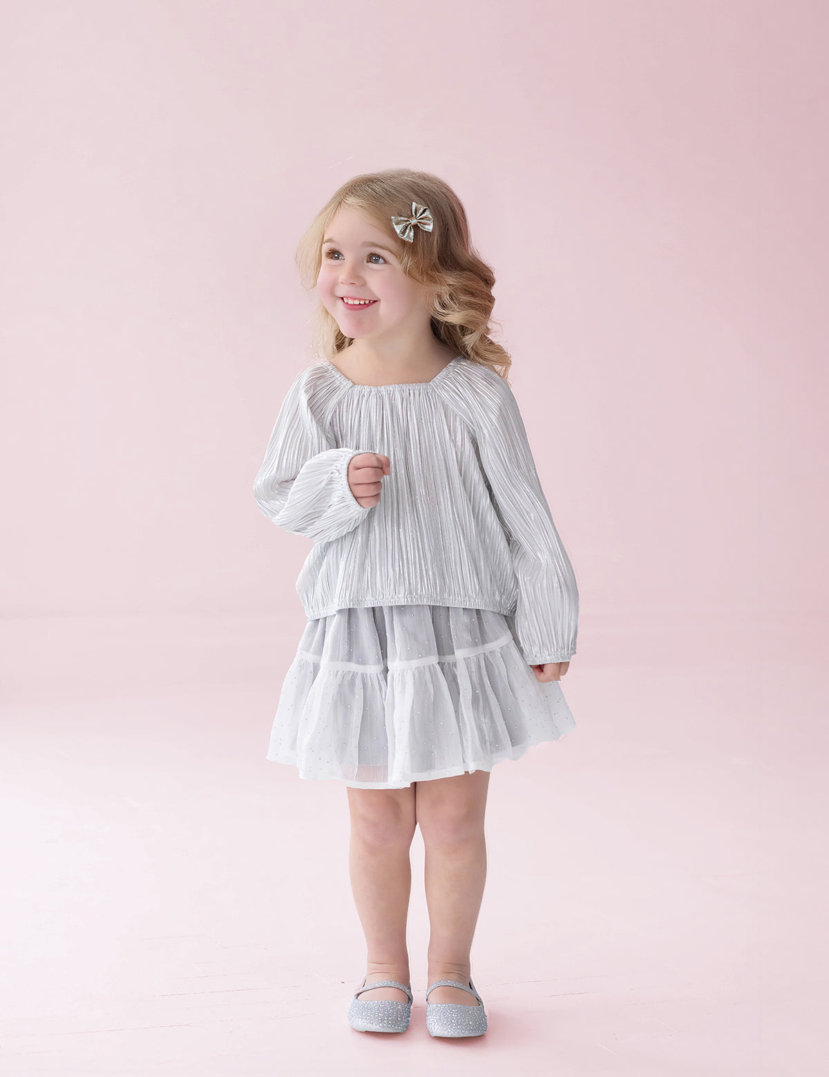 A young girl with blonde hair is wearing the Isobella and Chloe Shimmer Shine Long Sleeve Metallic Pleated Top and Tiered Skirt Set, styled with gray shoes, as she stands smiling against a pale pink background—an adorable choice for a girls party outfit.