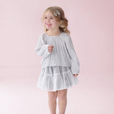 A young girl with blonde hair is wearing the Isobella and Chloe Shimmer Shine Long Sleeve Metallic Pleated Top and Tiered Skirt Set, styled with gray shoes, as she stands smiling against a pale pink background—an adorable choice for a girls party outfit.