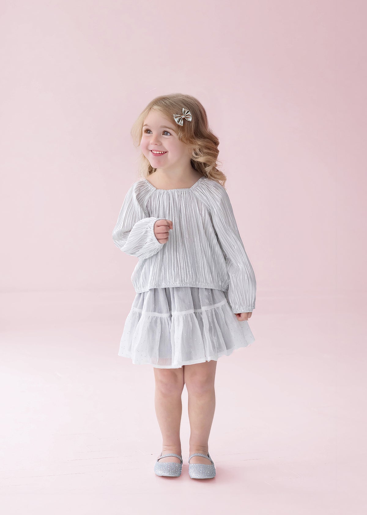 A young girl with blonde hair is wearing the Isobella and Chloe Shimmer Shine Long Sleeve Metallic Pleated Top and Tiered Skirt Set, styled with gray shoes, as she stands smiling against a pale pink background—an adorable choice for a girls party outfit.