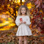 A young girl, dressed in the Mabel and Honey Autumn Breeze Long Sleeve Floral Ruffle Neck Top and Skirt Set, stands beneath autumn trees, smiling as she holds a red leaf. The crinkle cotton ensemble is paired perfectly with her pink boots, surrounded by vibrant fall foliage and scattered leaves on the ground.