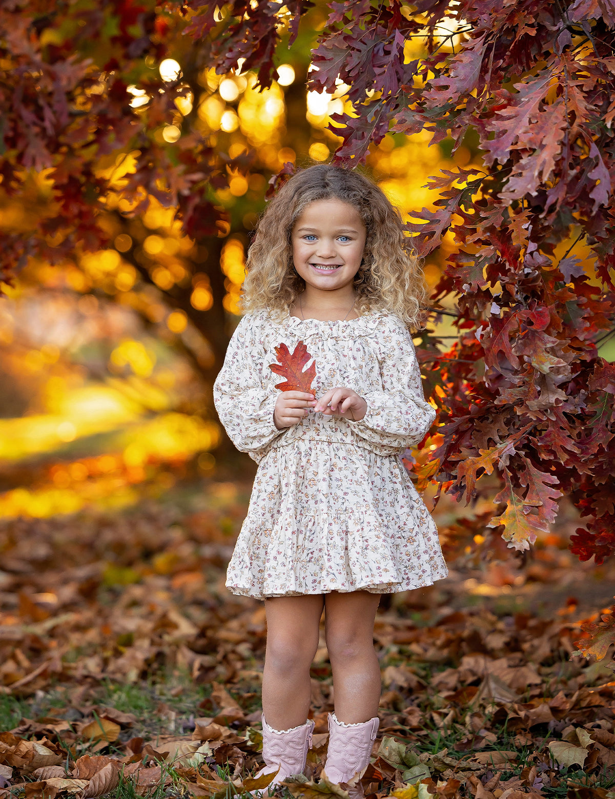 A young girl, dressed in the Mabel and Honey Autumn Breeze Long Sleeve Floral Ruffle Neck Top and Skirt Set, stands beneath autumn trees, smiling as she holds a red leaf. The crinkle cotton ensemble is paired perfectly with her pink boots, surrounded by vibrant fall foliage and scattered leaves on the ground.