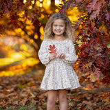 A young girl, dressed in the Mabel and Honey Autumn Breeze Long Sleeve Floral Ruffle Neck Top and Skirt Set, stands beneath autumn trees, smiling as she holds a red leaf. The crinkle cotton ensemble is paired perfectly with her pink boots, surrounded by vibrant fall foliage and scattered leaves on the ground.