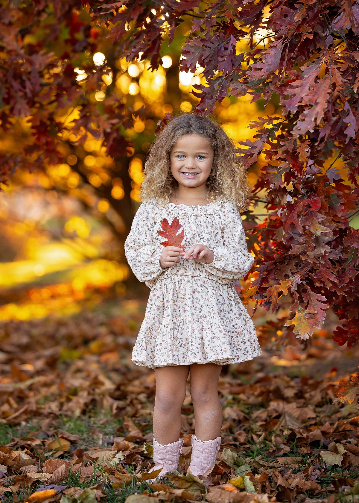 A young girl, dressed in the Mabel and Honey Autumn Breeze Long Sleeve Floral Ruffle Neck Top and Skirt Set, stands beneath autumn trees, smiling as she holds a red leaf. The crinkle cotton ensemble is paired perfectly with her pink boots, surrounded by vibrant fall foliage and scattered leaves on the ground.