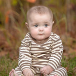A baby with short hair sits on the grass, looking ahead with a neutral expression, dressed in the Beckett and Bear Max Long Sleeve Striped Ribbed Trim Jogger Leg Romper featuring convenient snap closures. The background is blurred with autumnal colors.