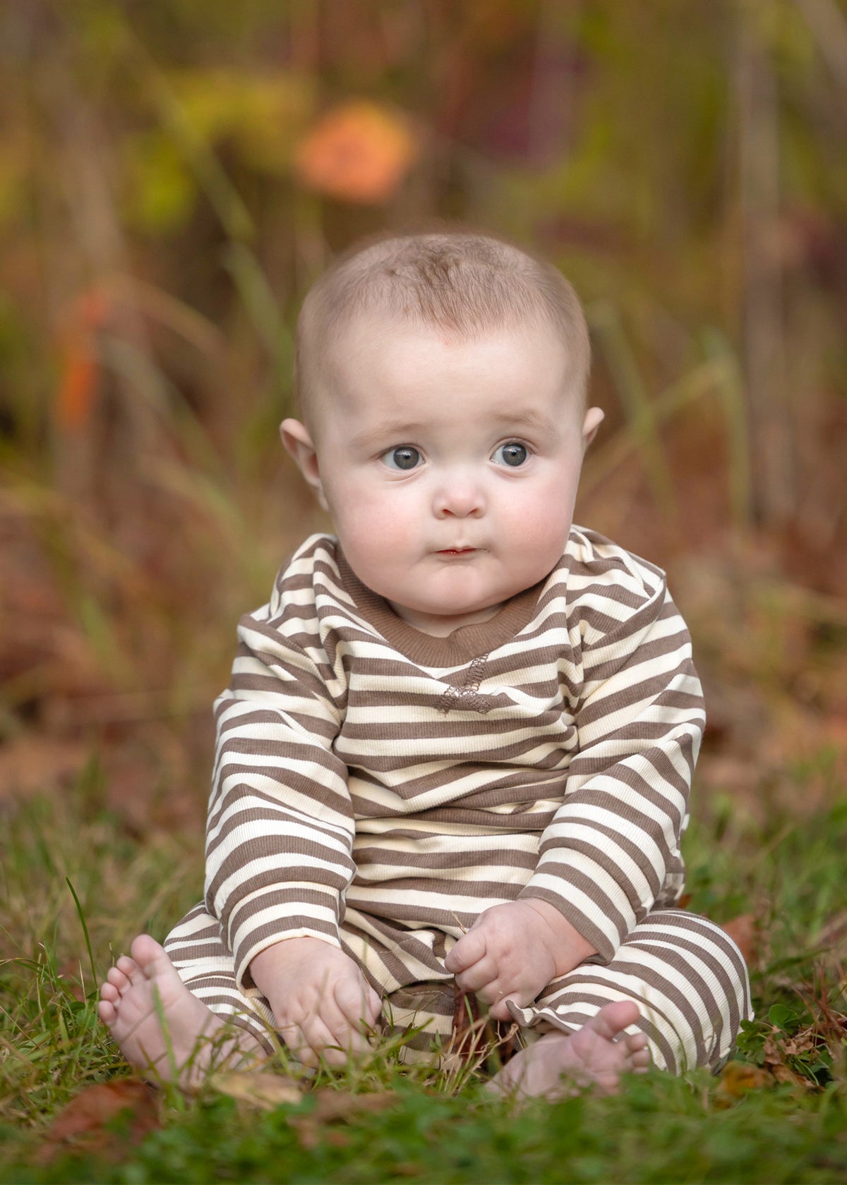 A baby with short hair sits on the grass, looking ahead with a neutral expression, dressed in the Beckett and Bear Max Long Sleeve Striped Ribbed Trim Jogger Leg Romper featuring convenient snap closures. The background is blurred with autumnal colors.