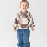 A young boy smiles while standing against a plain white background. He wears the Beckett and Bear Max Long Sleeve Striped Raglan Ribbed Crew Neck Top—a brown and white striped piece crafted from soft cotton, perfect for fall layering—paired with blue pants with elastic cuffs and white sneakers featuring blue and red accents.