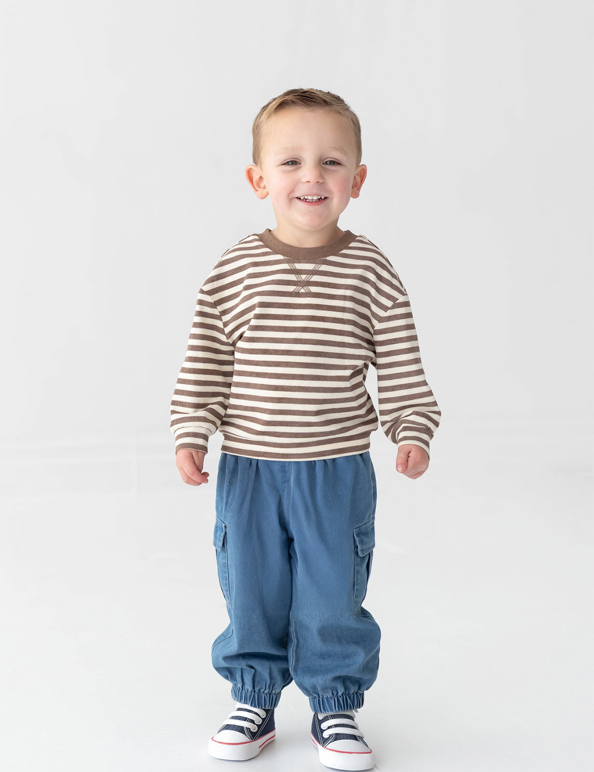 A young boy smiles while standing against a plain white background. He wears the Beckett and Bear Max Long Sleeve Striped Raglan Ribbed Crew Neck Top—a brown and white striped piece crafted from soft cotton, perfect for fall layering—paired with blue pants with elastic cuffs and white sneakers featuring blue and red accents.