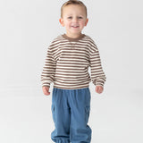 A young boy smiles while standing against a plain white background. He wears the Beckett and Bear Max Long Sleeve Striped Raglan Ribbed Crew Neck Top—a brown and white striped piece crafted from soft cotton, perfect for fall layering—paired with blue pants with elastic cuffs and white sneakers featuring blue and red accents.