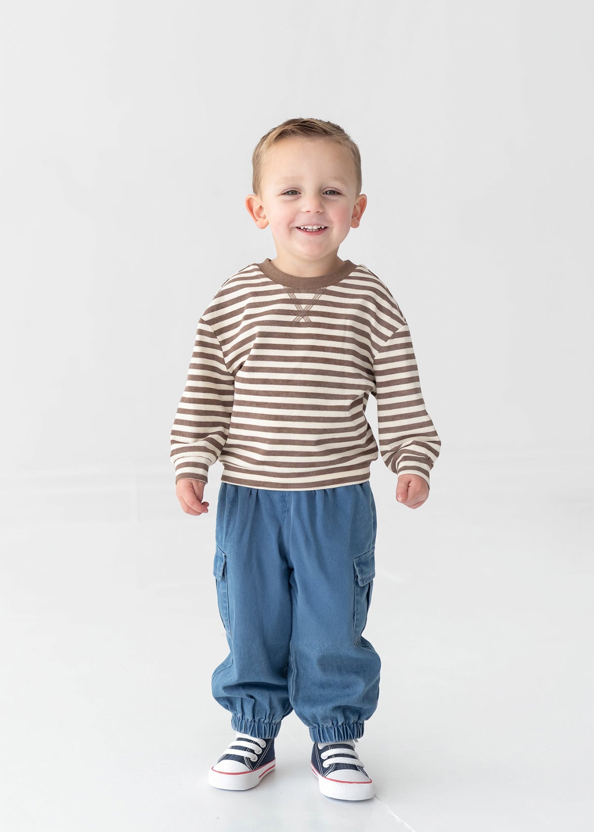 A young boy smiles while standing against a plain white background. He wears the Beckett and Bear Max Long Sleeve Striped Raglan Ribbed Crew Neck Top—a brown and white striped piece crafted from soft cotton, perfect for fall layering—paired with blue pants with elastic cuffs and white sneakers featuring blue and red accents.