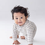 A smiling baby with curly hair is crawling on a white background, dressed in the Beckett and Bear Ethan Long Sleeve Striped Ribbed Trim Straight Leg Romper, featuring gray and white stripes, soft ribbed cotton fabric, and convenient snap closures.