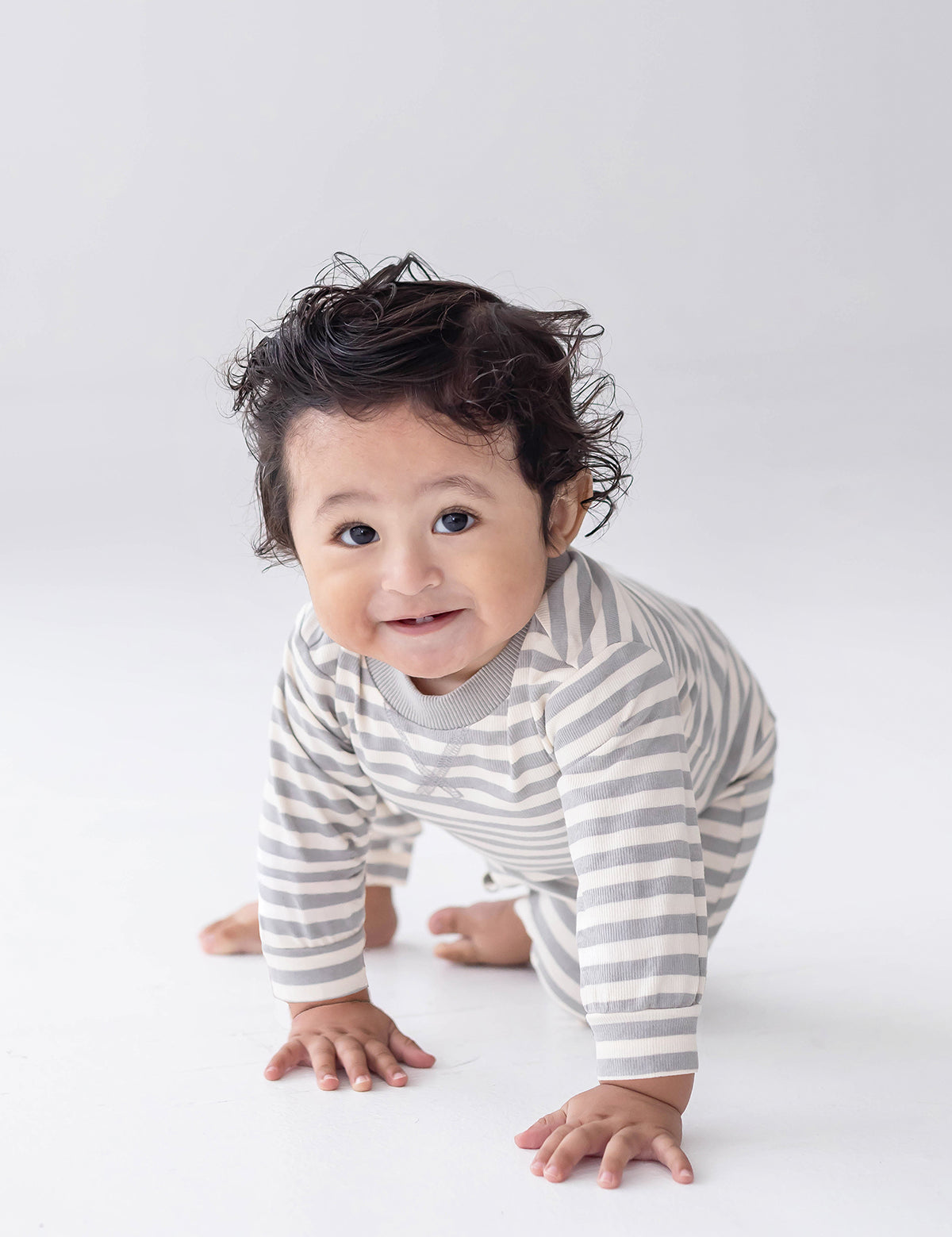 A smiling baby with curly hair is crawling on a white background, dressed in the Beckett and Bear Ethan Long Sleeve Striped Ribbed Trim Straight Leg Romper, featuring gray and white stripes, soft ribbed cotton fabric, and convenient snap closures.