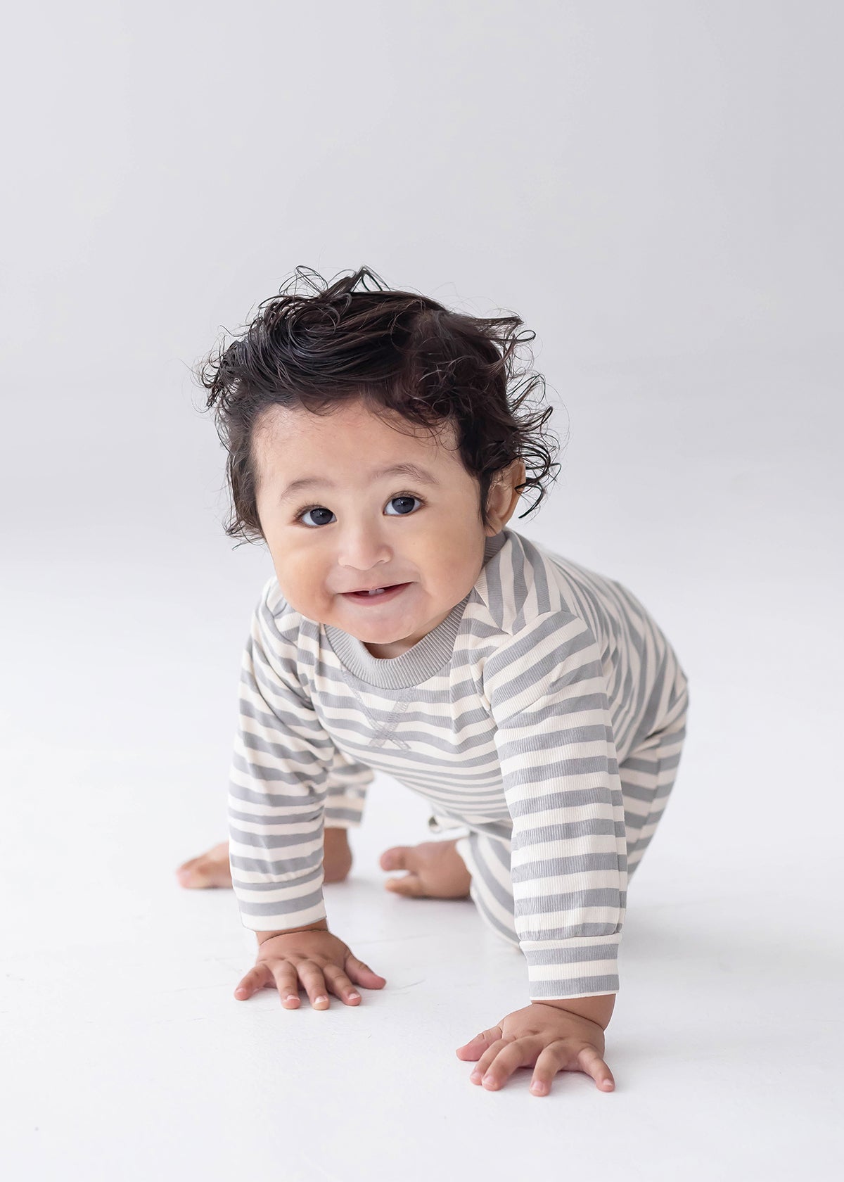 A smiling baby with curly hair is crawling on a white background, dressed in the Beckett and Bear Ethan Long Sleeve Striped Ribbed Trim Straight Leg Romper, featuring gray and white stripes, soft ribbed cotton fabric, and convenient snap closures.
