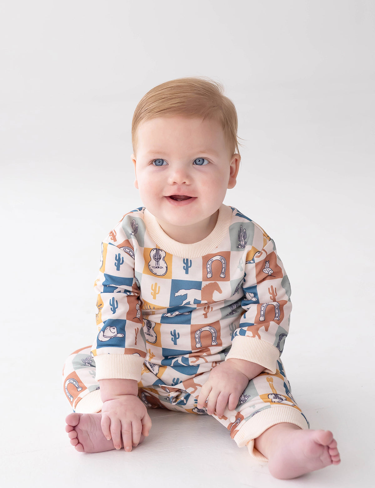 A baby with light hair and blue eyes sits on a white surface, smiling and looking slightly off-camera while wearing the Beckett and Bear Westin Long Sleeve Western Print Straight Leg Romper. Crafted from soft baby cotton knit, this matching outfit features vibrant western-themed patterns including cacti, horseshoes, cowboy hats, and a playful checkerboard print.