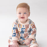 A baby with light hair and blue eyes sits on a white surface, smiling and looking slightly off-camera while wearing the Beckett and Bear Westin Long Sleeve Western Print Straight Leg Romper. Crafted from soft baby cotton knit, this matching outfit features vibrant western-themed patterns including cacti, horseshoes, cowboy hats, and a playful checkerboard print.