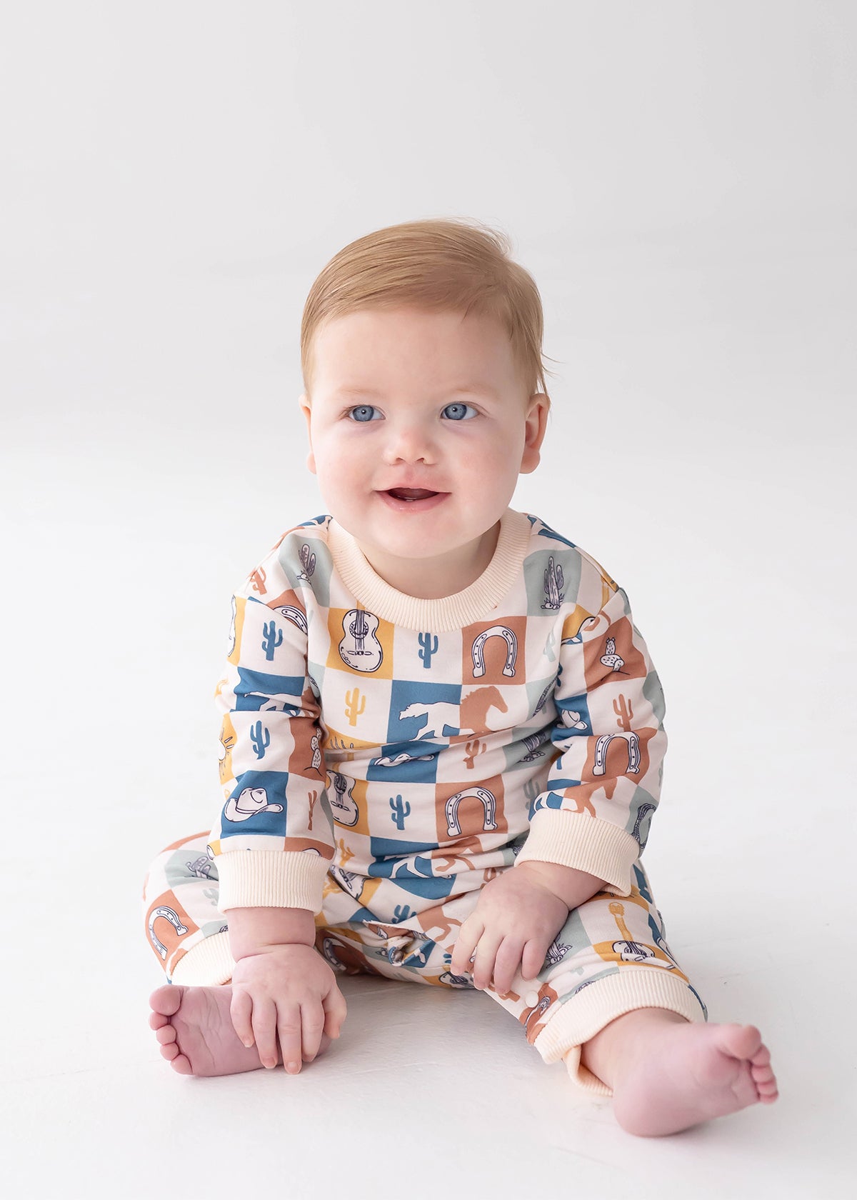 A baby with light hair and blue eyes sits on a white surface, smiling and looking slightly off-camera while wearing the Beckett and Bear Westin Long Sleeve Western Print Straight Leg Romper. Crafted from soft baby cotton knit, this matching outfit features vibrant western-themed patterns including cacti, horseshoes, cowboy hats, and a playful checkerboard print.