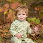 A smiling toddler with light brown hair sits on the grass, wearing the Beckett and Bear Matthew Long Sleeve Fish Print Ribbed Trim Button Placket Romper. Autumn leaves and plants are softly blurred in the background as the child waves with one hand.