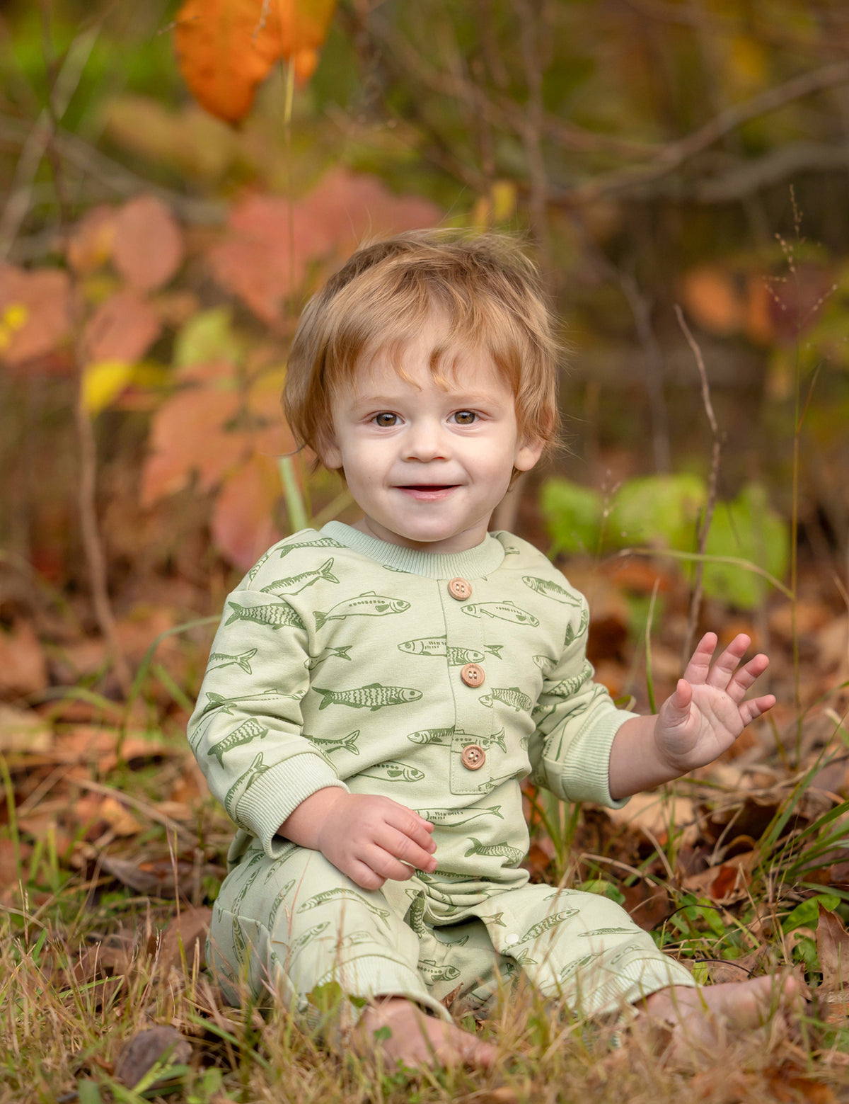 A smiling toddler with light brown hair sits on the grass, wearing the Beckett and Bear Matthew Long Sleeve Fish Print Ribbed Trim Button Placket Romper. Autumn leaves and plants are softly blurred in the background as the child waves with one hand.