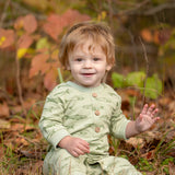 A smiling toddler with light brown hair sits on the grass, wearing the Beckett and Bear Matthew Long Sleeve Fish Print Ribbed Trim Button Placket Romper. Autumn leaves and plants are softly blurred in the background as the child waves with one hand.