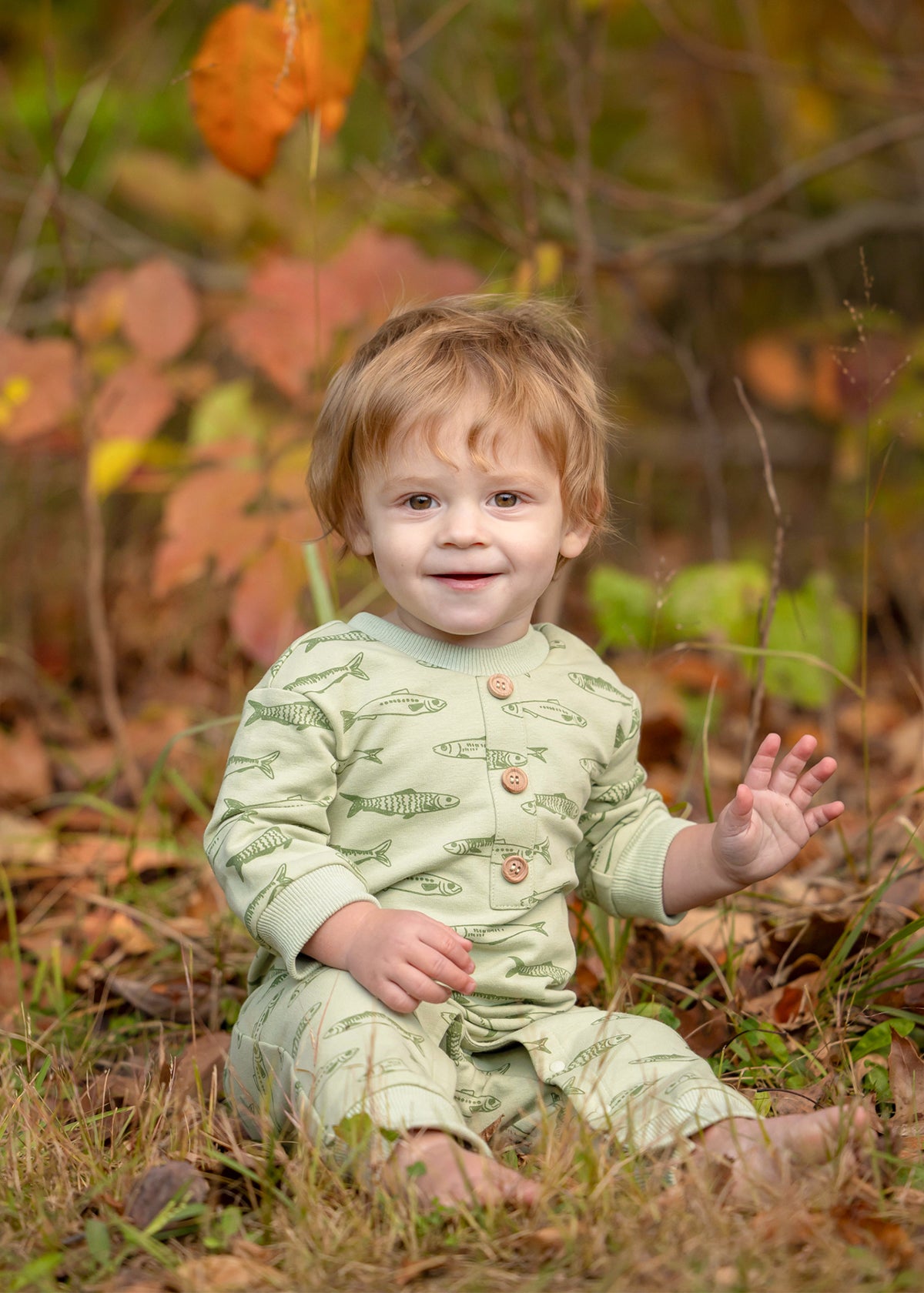 A smiling toddler with light brown hair sits on the grass, wearing the Beckett and Bear Matthew Long Sleeve Fish Print Ribbed Trim Button Placket Romper. Autumn leaves and plants are softly blurred in the background as the child waves with one hand.
