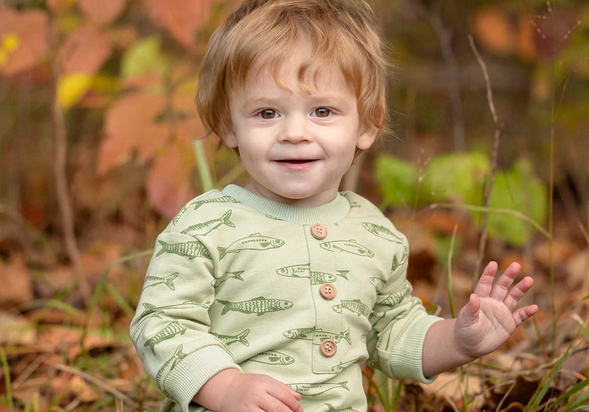A smiling toddler with light brown hair sits on the grass, wearing the Beckett and Bear Matthew Long Sleeve Fish Print Ribbed Trim Button Placket Romper. Autumn leaves and plants are softly blurred in the background as the child waves with one hand.