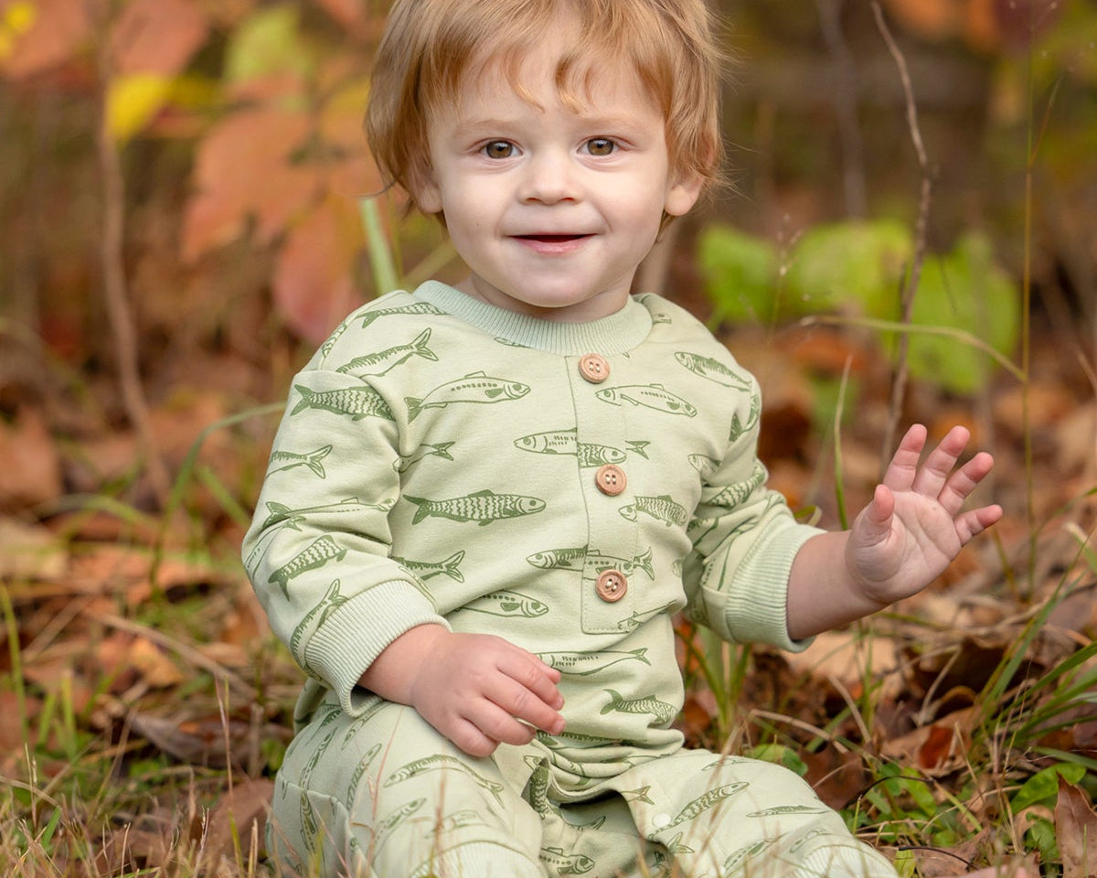 A smiling toddler with light brown hair sits on the grass, wearing the Beckett and Bear Matthew Long Sleeve Fish Print Ribbed Trim Button Placket Romper. Autumn leaves and plants are softly blurred in the background as the child waves with one hand.