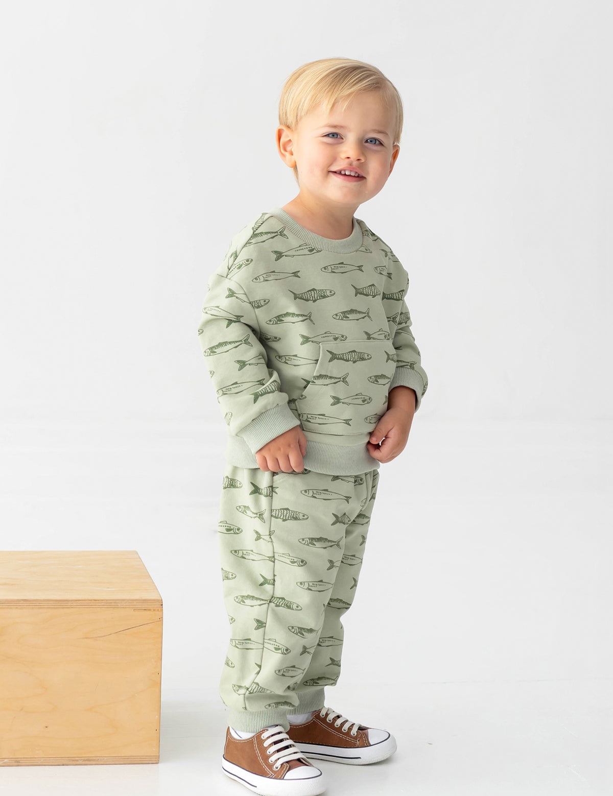 A smiling blond-haired toddler wears the Beckett and Bear Matthew Long Sleeve Fish Print Sweatshirt and Jogger Set in sage green, made from a soft cotton blend fleece, along with brown sneakers, and stands beside a wooden block against a plain white background.
