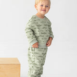 A smiling blond-haired toddler wears the Beckett and Bear Matthew Long Sleeve Fish Print Sweatshirt and Jogger Set in sage green, made from a soft cotton blend fleece, along with brown sneakers, and stands beside a wooden block against a plain white background.